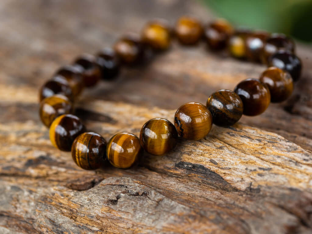 L'image présente un bracelet en pierre avec des perles d’œil de tigre, avec des nuances brunes et dorées, reposant sur un fond en bois naturel. Ce bijou évoque élégance et force.